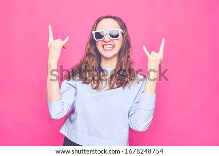 Stylish caucasian girl in a pale blue t-shirt and 8-bit glasses on a pink background. A two-handed gesture is rock. Close up. Toned.