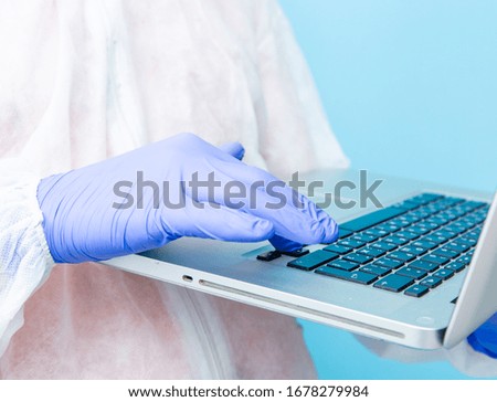 A virologist in a white mask and blue rubber gloves is dressed in a viral protective suit holding a gray laptop in his hands and looking at the information in it