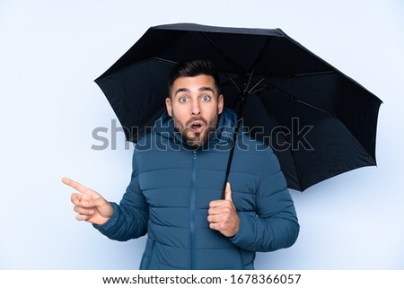 Man holding an umbrella over isolated background surprised and pointing side