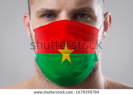 Young man with sore eyes in a medical mask painted in the colors of the national flag of Burkino Faso
 Medical protection against airborne diseases, coronavirus. Man is afraid of getting the flu