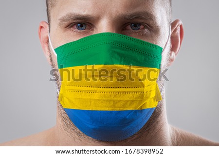 Young man with sore eyes in a medical mask painted in the colors of the national flag of Gabon. Medical protection against airborne diseases, coronavirus. Man is afraid of getting the flu