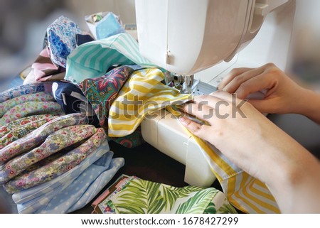 Bright, closeup shot of ASEAN woman hands using sewing machine to make 3-layer cloth face masks in different colorful patterns to support demands during COVID-19 pandemic and shortage of sanitary mask