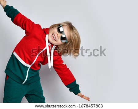 Portrait of happy active frolic blond kid boy in dark gray and red hoodie, pants and sunglasses is playing aircraft pilot with his arms spread on gray background with copy space