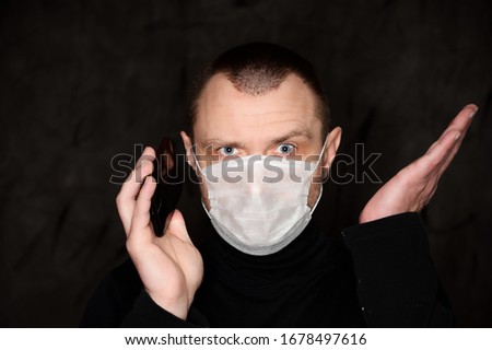 Close-up portrait of an adult man with emotions in a medical mask of a Caucasian on a black background with a phone in his hands
