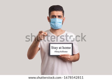 #StayAtHome. Man in protective mask holding Stay at home inscription and pointing to camera, warning of coronavirus quarantine, Covid-19 epidemic, preventive measures for spread of infectious disease. Royalty-Free Stock Photo #1678542811