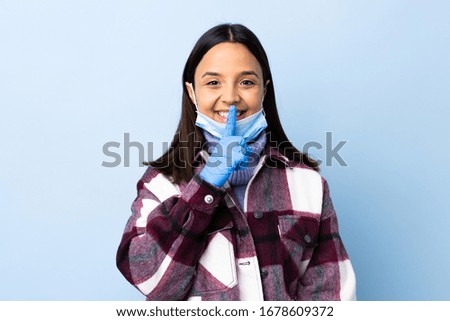 Young brunette mixed race woman protecting from the coronavirus with a mask and gloves over isolated blue background showing a sign of silence gesture putting finger in mouth
