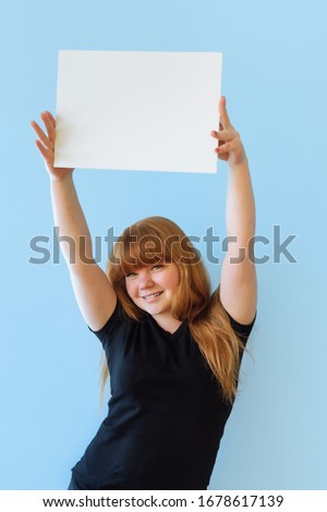 young woman of Caucasian appearance with red hair holds a poster with copyspacer with two hands, she smiles, on an isolated blue background, dressed in black casual clothes