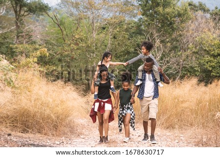 Happy big mixed race family, father and mother carrying our daughter walking along yellow field to campsite. Travel and hiking in vacation concept.