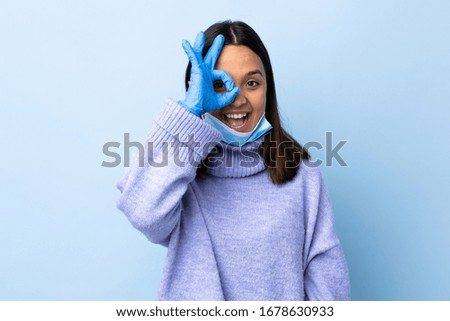 Young brunette mixed race woman protecting from the coronavirus with a mask and gloves over isolated blue background showing ok sign with fingers