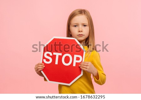 Portrait of cute little girl holding Stop symbol, red traffic sign and looking with serious responsible expression, warning about road safety rules. indoor studio shot isolated on pink background