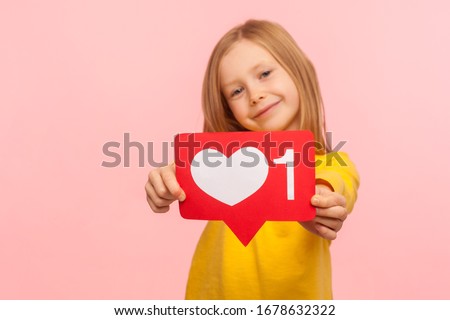 Child blog, trendy content. Portrait of adorable cheerful little girl showing heart like icon of social media and looking at camera with cute smile. indoor studio shot isolated on pink background