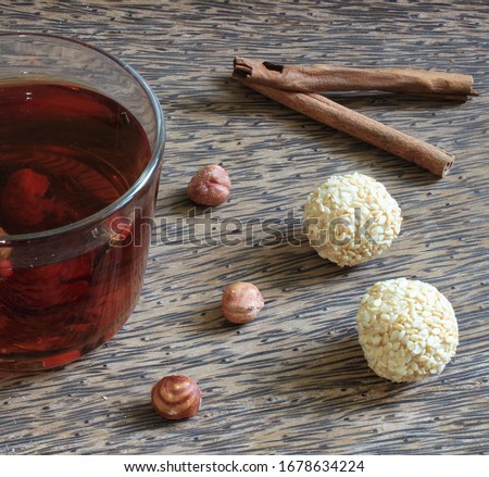 Glass cup of black tea with cinnamon, huzelnuts and two white round sweets on the wooden table without people. Brown color new peaceful wallpaper 