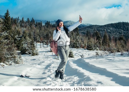 A girl is taking pictures of a mountain landscape. Beautiful mountain lake in winter. Snowy mountains and trees. Outdoor activities. Red jacket and backpack.