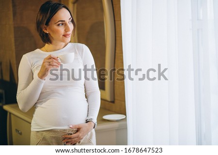 Young pregnant woman standing by the window and drinkig coffee