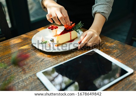 Waitress preparing cheesecake with berries on worktop, looking at recipe on tablet screen.