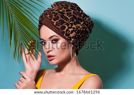 An attractive young woman in a stylish turban made of leopard print fabric on a blue background. Girl with bright makeup. Beach style, palm branch and heat