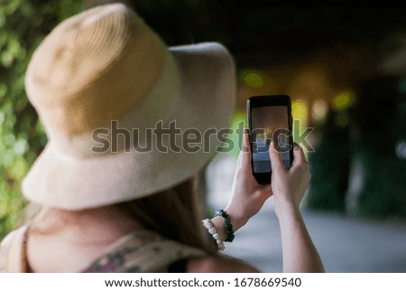Girl with hat taking photos of the park road using the phone. Close-up shot of the hand holding the phone with the columns arch behind.