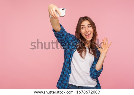 Communication on mobile phone. Portrait of cute cheerful girl in checkered shirt smiling and gesturing hello while having positive talk on video call, online chatting. indoor studio shot, isolated