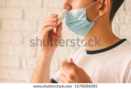 Closeup of young man with sickness using nasal spray for his congested nose. Confident ill doctor in protective medical mask on face using nose drops for runny nose. Coronavirus COVID-19 symptoms Royalty-Free Stock Photo #1678686085