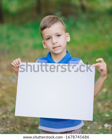 Closeup view vertical photography of cute little kid standing outdoor in green summer nature background and holding big white empty blank paper in hands.