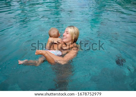 Beautiful mother teaching cute baby girl how to swim in a swimming pool. Child having fun in water with mom.