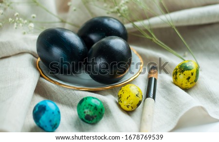 black eggs on a plate with a gold border and next to it multicolored quail eggs on a gray linen background. horizontal photo