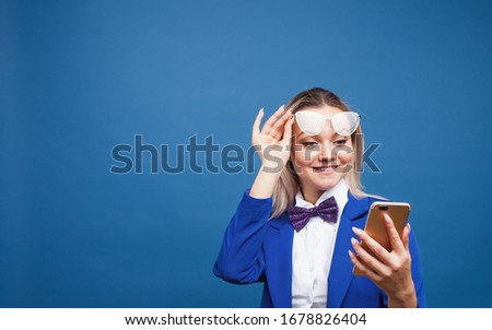 Cute and funny businesswoman in a stylish blue jacket and bow tie uses a smartphone. Girl in suit with mobile phone happily laughs, copy space