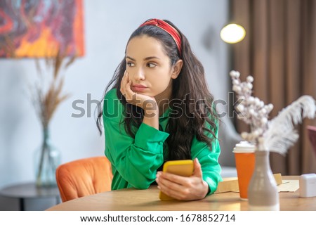 Moment of waiting. Beautiful young woman with a yellow smartphone in hand, sitting at a table in anticipation, propping her head with her hand, looking to the side.