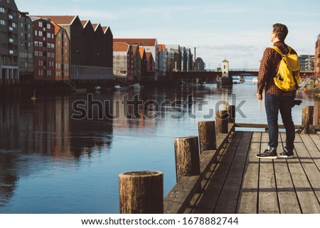 Photographer with yellow backpack standing on wooden pier taking photo on against city and river background