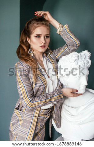 a blonde girl in a beige yellow checked suit with a skirt in high heels with her hair in a ponytail poses on an emerald background in the background a plaster sculpture of a head in a photo Studio