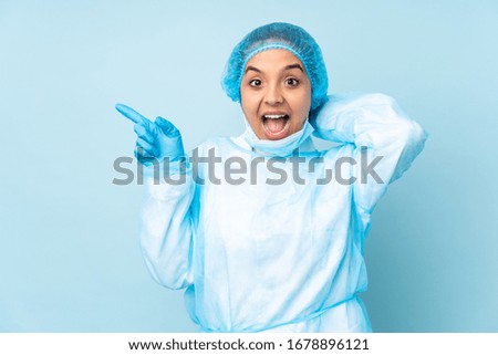 Young surgeon Indian woman in blue uniform surprised and pointing finger to the side