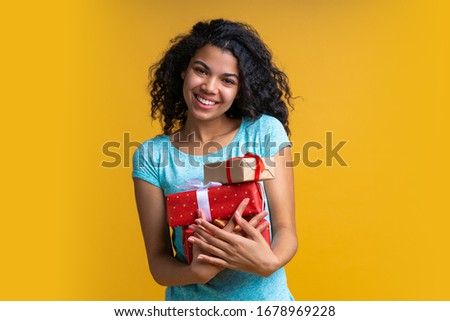 Portrait of smiling cute casually dressed dark skinned girl posing with a bunch of gift boxes in hands isolated over bright colored yellow background.