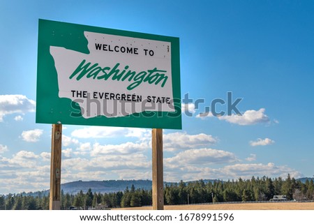A roadside sign with welcome to Washington State, the evergreen state written on it with a rural setting and blue sky behind.