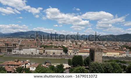 View of an Italian city with a tower and mountains under a blue sky in the clouds