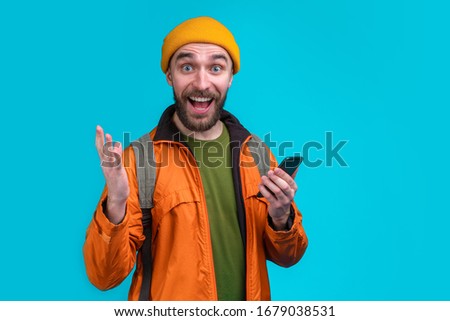 Half length studio portrait of casually dressed male tourist with a backpack  holding mobile phone in hand with happy excited mouth open face expresson. Isolated over blue background.