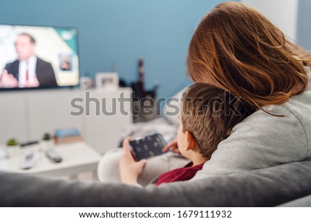 Mother and son young woman and her boy child caucasian kid sitting on sofa bed at home making video call or playing video games on mobile smart phone and watch tv during quarantine lockdown back view
