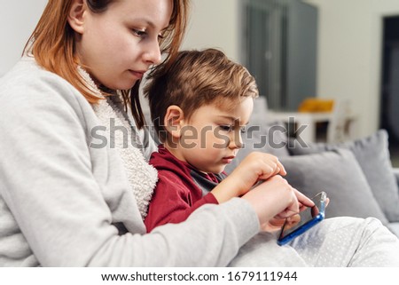 Mother and son young woman and her boy child caucasian kid sitting on the sofa bed at home making video call or playing video games on mobile smart phone during quarantine lockdown