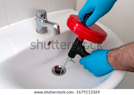 Plumbing issues, occupation in sanitation and handyman contractor concept with plumber repairing drain with plumbers snake (steel spiral that twists through pipes to collect dirt) in residential sink Royalty-Free Stock Photo #1679113369