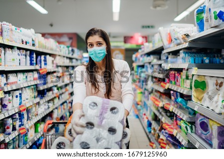 Woman shopper with mask and gloves panic buying and hoarding toilette paper in supply store.Preparing for pathogen virus pandemic quarantine.Prepper buying bulk cleaning supplies due to Covid-19. Royalty-Free Stock Photo #1679125960