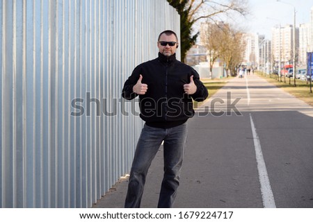 Street photo of a handsome man in a black jacket with glasses in the city on a sunny day near the wall shows his fingers to the sides