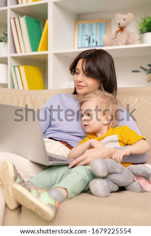 Blond-haired boy embraced by mom sitting on sofa and watching curious cartoon on laptop at home