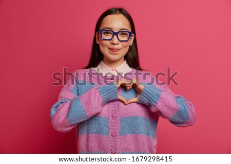 Pleasant looking tender woman confesses in love, makes heart sign, likes someone, expresses good feelings, wears transparent spectacles and striped jumper, shows sympathy, has romantic relationship