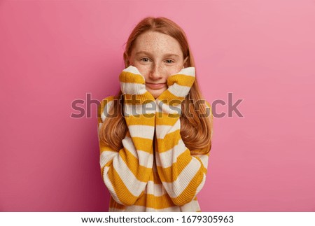 Portrait of pleasant looking ginger European girl has freckled face, keeps hands under chin, looks calm and satisfied, wears striped jumper, being very obedient, isolated on pink background.