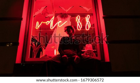 Fashion tomboy teen hipster short hairstyle gen z girl wear stylish clothes glasses sit on red street window near neon sign. Female model woman relax in city night light 80s 90s style glow. Royalty-Free Stock Photo #1679306572