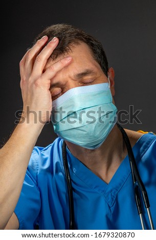 Portrait of a worried male doctor with hand on head and eyes closed, wearing blue hospital scrubs ,stethoscope with surgical face mask, against a dark background.
