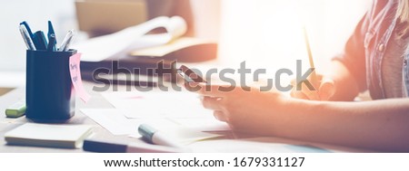 Woman working in open space office. Paperwork on the table, mobile phone. Sun glare, film effect and blurry background. Wide screen. Panoramic