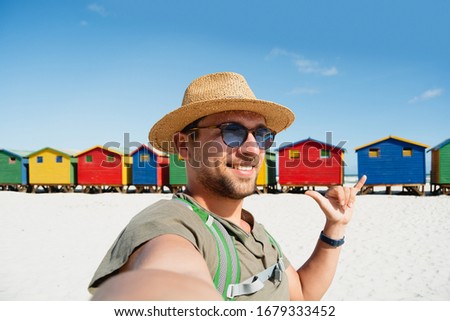 Traveller millennial guy in straw hat taking selfie picture with colourful wooden beach huts on white sand beach in Simon Town, South Africa. Sunny summer holiday vacation relax time at seaside.