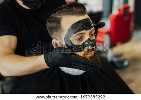 Barber applies black charcoal mask to man face to clean pore skin and remove acne from nose in barbershop. Close Up of handsome serious man with black mask against dots on face sitting on chair. Royalty-Free Stock Photo #1679385292