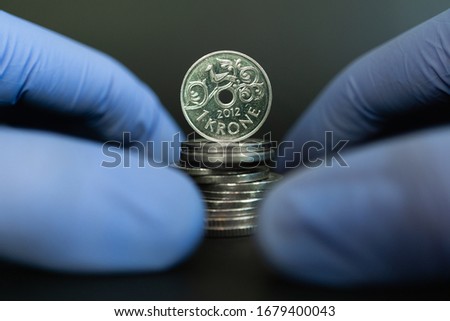 Norwegian financial crisis and economy during coronavirus or covid-19 pandemic. One Norwegian krone (NOK) coin standing on stack of coins surrounded by hands with protective gloves. Economy of Norway.