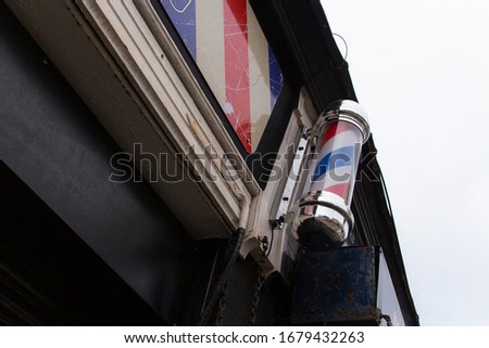 Front entrance of a barber shop, with the traditional red, blue and white pattern on its signage and store front. Barbers are hair cutters for men, or those with short hair. Electric shavers are used.